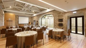 Un salón de conferencias con varias mesas redondas, suelo de madera, paredes de piedra y una pantalla grande para presentaciones.
