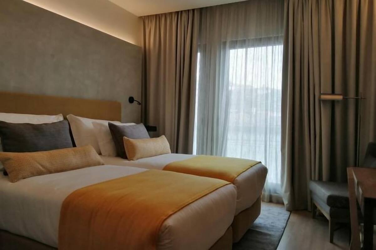 A hotel room with two beds, a desk, and a large window with curtains.