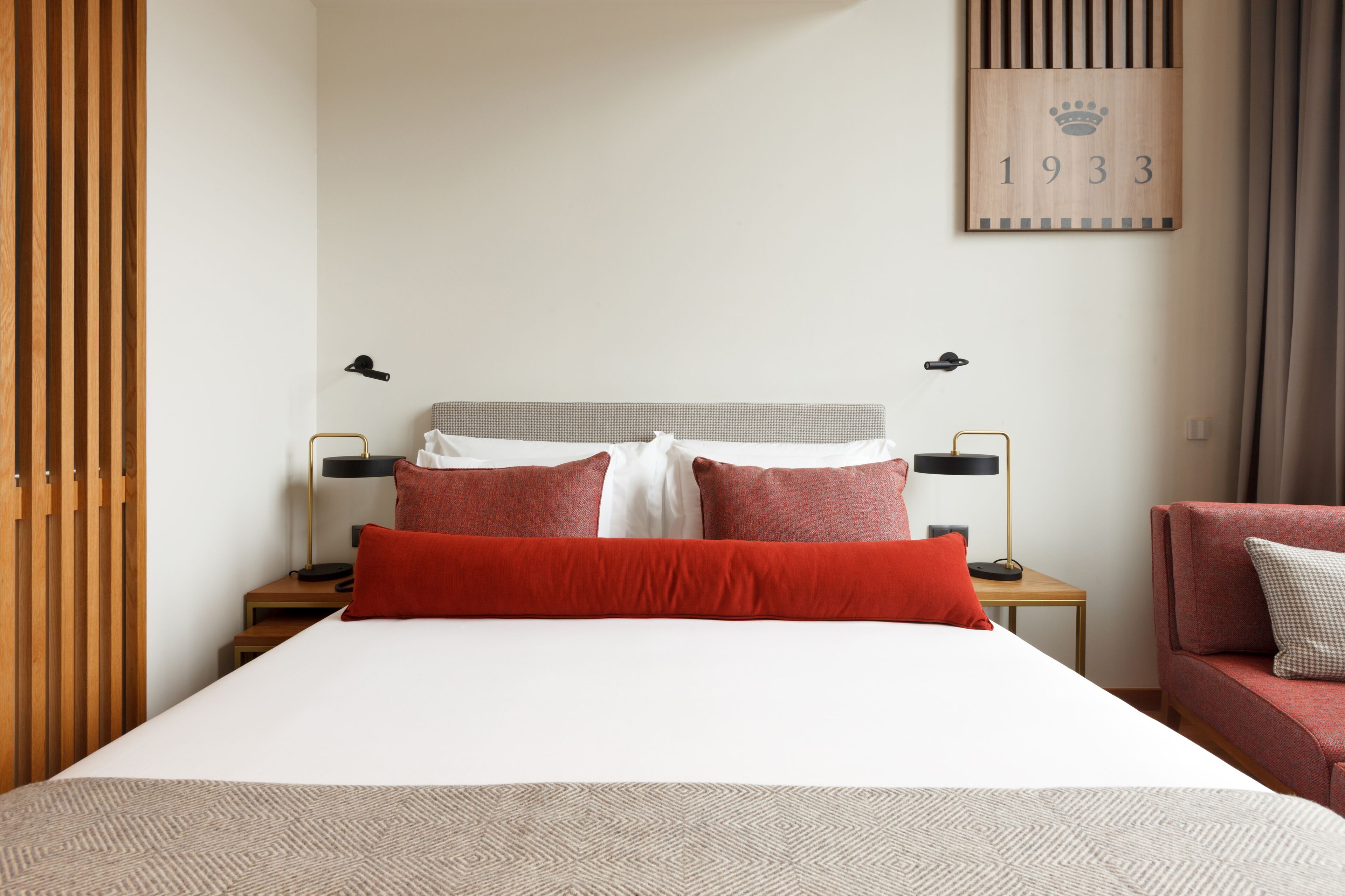 A neatly arranged hotel room with a large bed, two bedside tables, a red sofa, and a wall-mounted number sign.