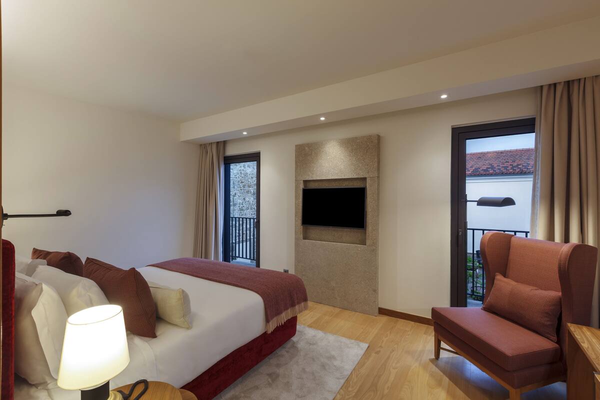 A modern hotel room with a bed, a sofa, a TV mounted on the wall, and a balcony with a view.