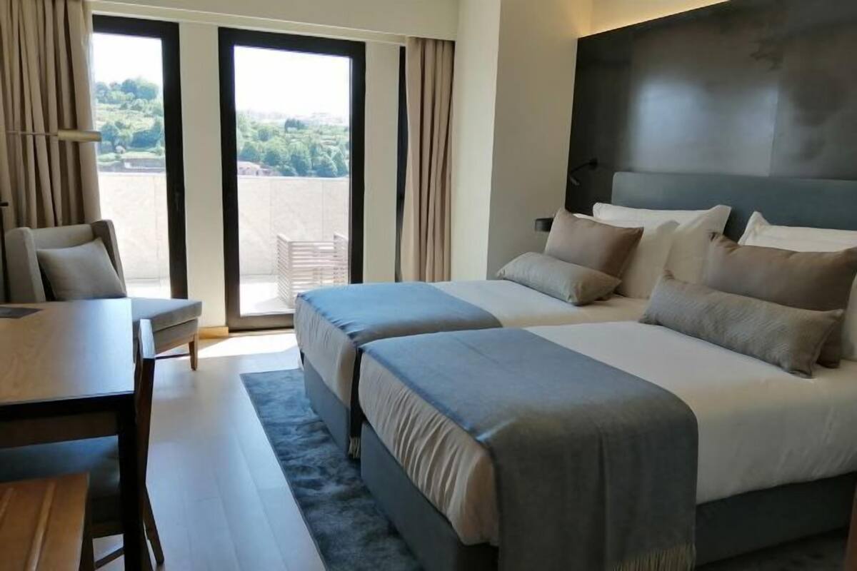 A hotel room with two beds, a desk, and a view of greenery through large windows.