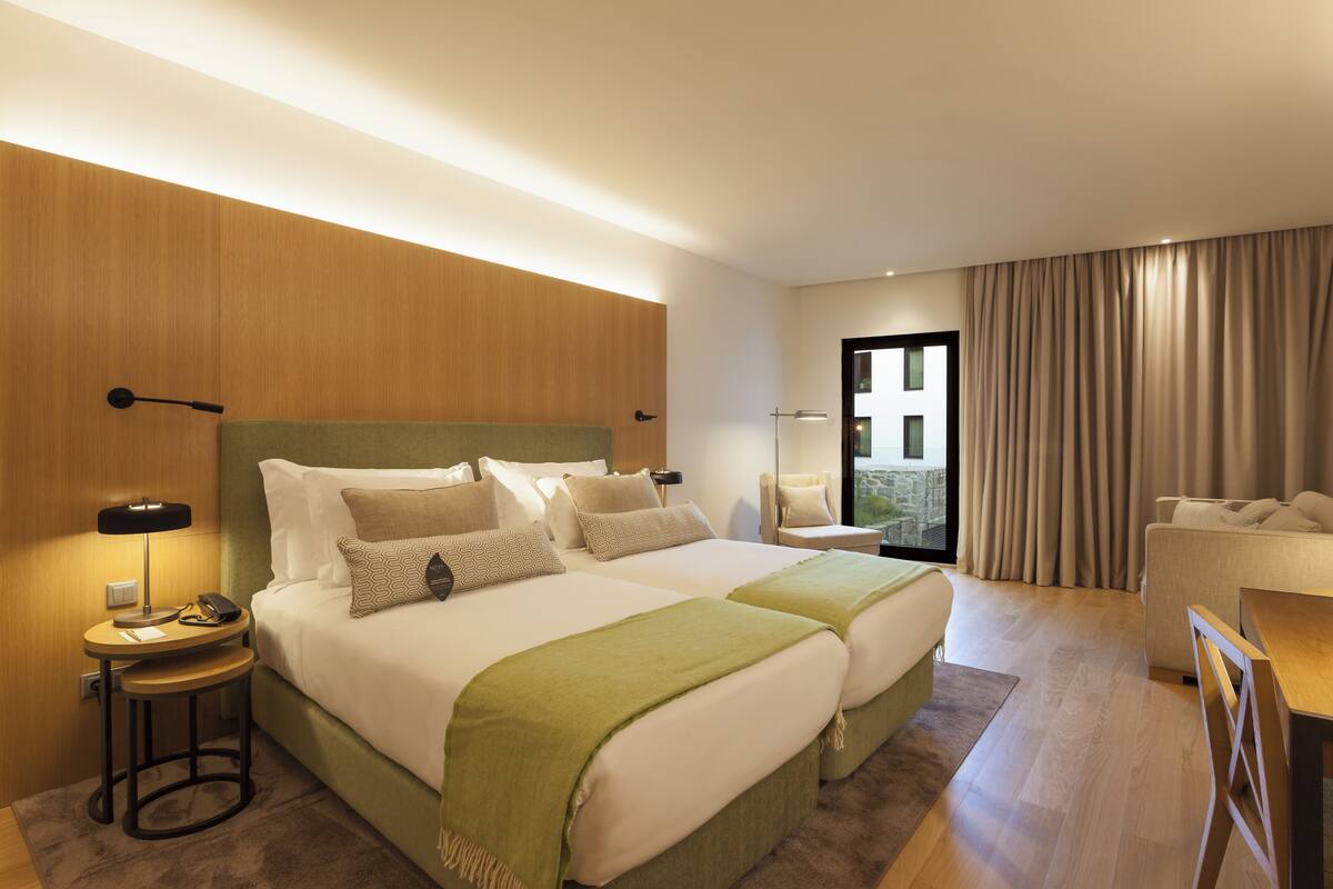 A hotel room with two beds, a desk, a chair, and a view of the outdoors.