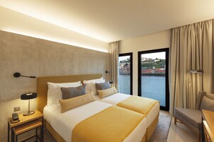 A hotel room with two beds, a desk, a chair, a lamp, and a view of a cityscape through the window.