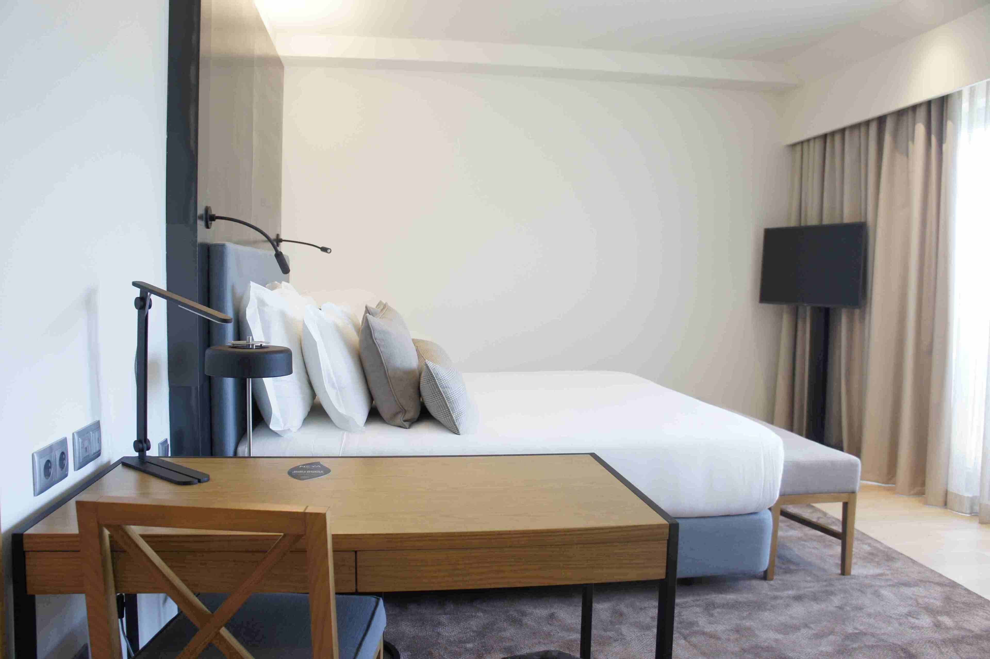 A modern hotel room with a large bed, a desk, a chair, a flat-screen TV, and a large window with curtains.