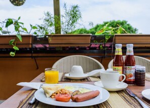 A breakfast table with toast, beans, sausage, tomato, and orange juice. There are two bottles of sauce and a cup of coffee. The table is outdoors with a wicker chair and greenery in the background. - Dafu Boutique Hotel Stonetown (Stonetown)