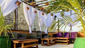Un espacio al aire libre con mesas y sillas de madera, plantas en macetas y un muro de piedra como fondo.