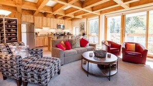 A spacious living area with a high ceiling, wooden beams, a kitchen with modern appliances, and large windows offering a view of the outdoors. - Treetops Condo Short Walk to Base of Eagle Bahn Gondola by RedAwning (Vail)