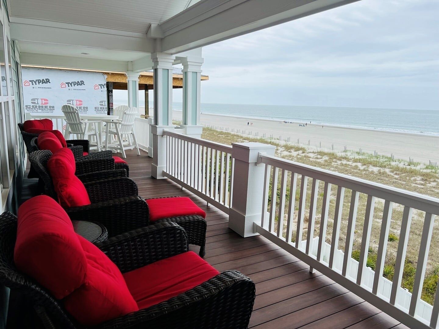 A covered outdoor seating area with wicker furniture and red cushions, overlooking a beach.