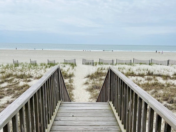 Ein hölzerner Steg führt zu einem Sandstrand mit freiem Blick auf das Meer.