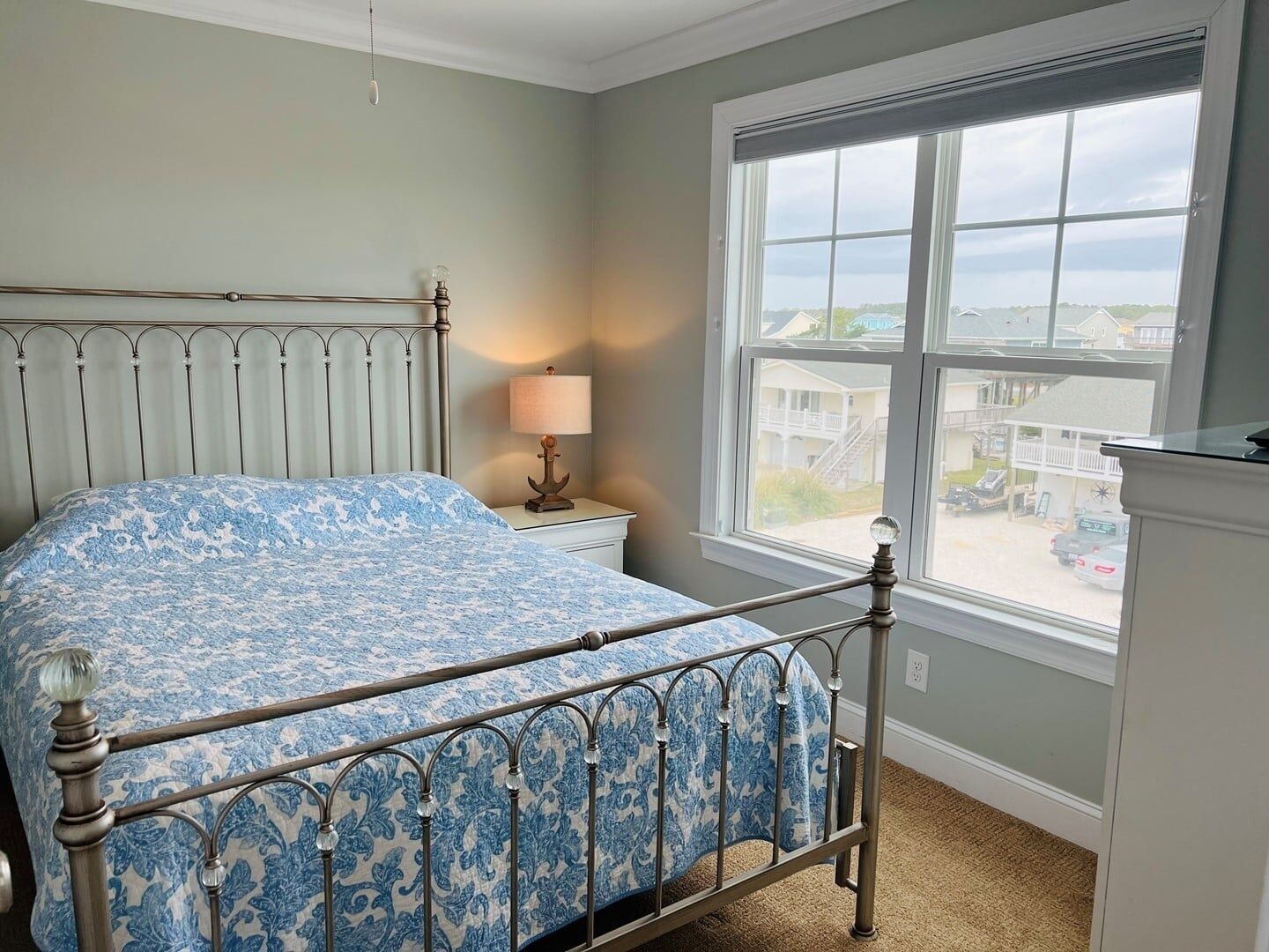 A bedroom with a bed, a nightstand, a lamp, and a window with a view of houses and cars.
