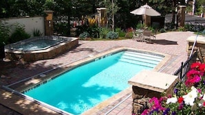 A rectangular swimming pool with a hot tub, surrounded by a brick patio and lush greenery.