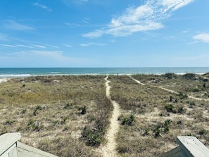 A beach path leading to the ocean with sparse vegetation and a clear sky.