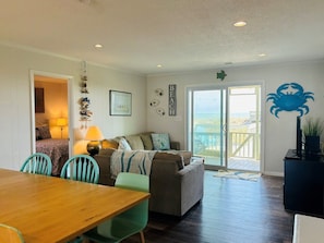 Relax in this open-plan living space with a dining table, sofa, and TV stand, leading to a balcony with a beach view.