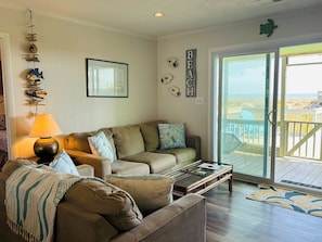 A living room with a sofa, a coffee table, a lamp, and a view of the beach through a sliding glass door.