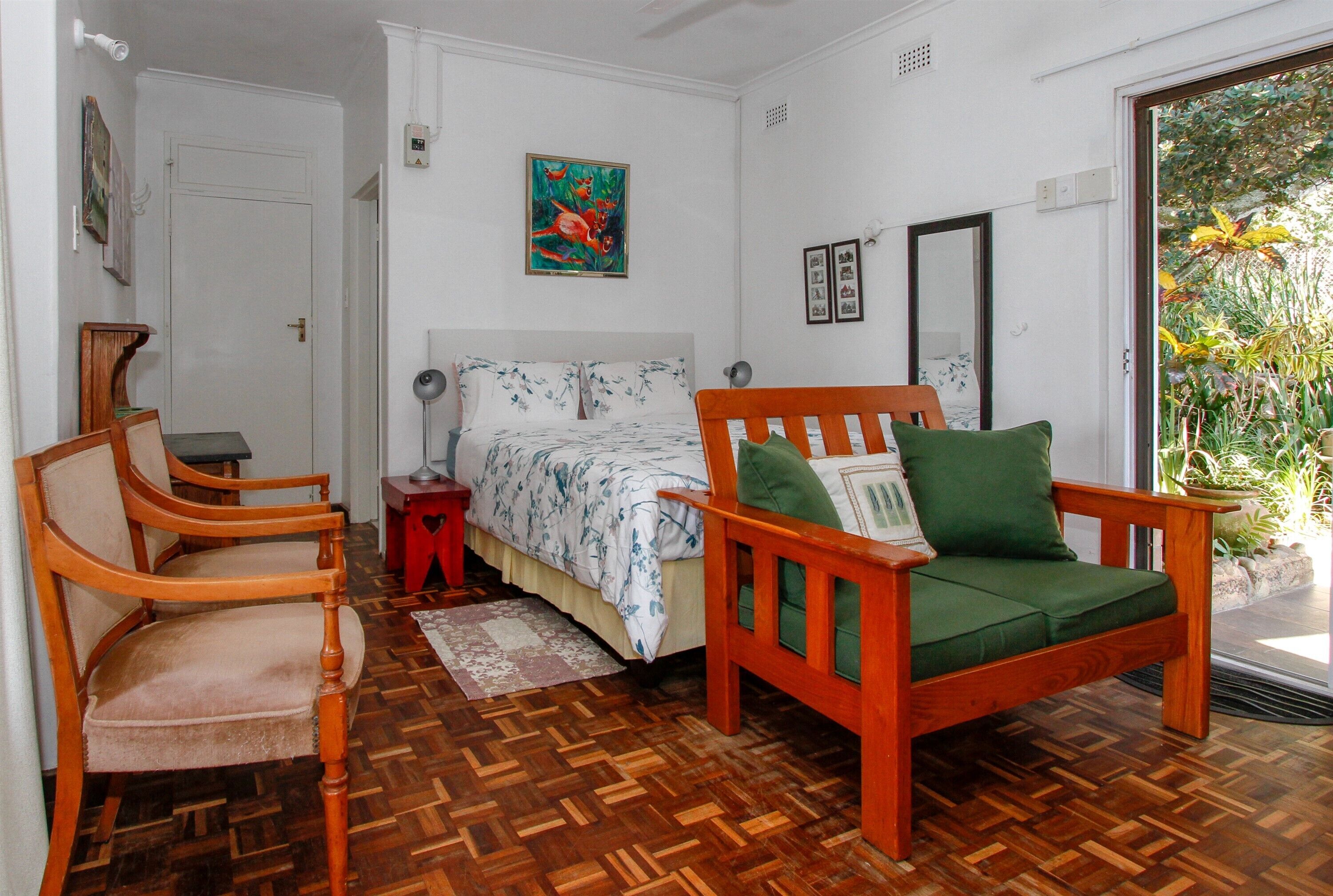 A room with a wooden floor, a bed with floral bedding, a green cushioned chair, a wooden chair, a small table, and a door.