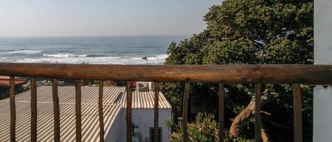 A balcony with a view of the ocean, a tree, and a building with a tiled roof.