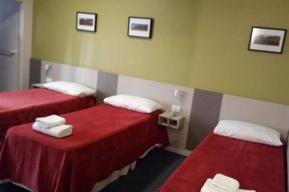 Une chambre d’hôtel avec trois lits simples, chacun recouvert d’une couverture rouge et agrémenté d’oreillers blancs. Deux tableaux encadrés sont accrochés au mur, au-dessus des lits.