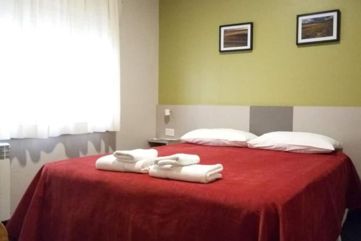 Une chambre d’hôtel avec une couverture de lit rouge, deux oreillers et une serviette blanche soigneusement pliée posée sur le lit.