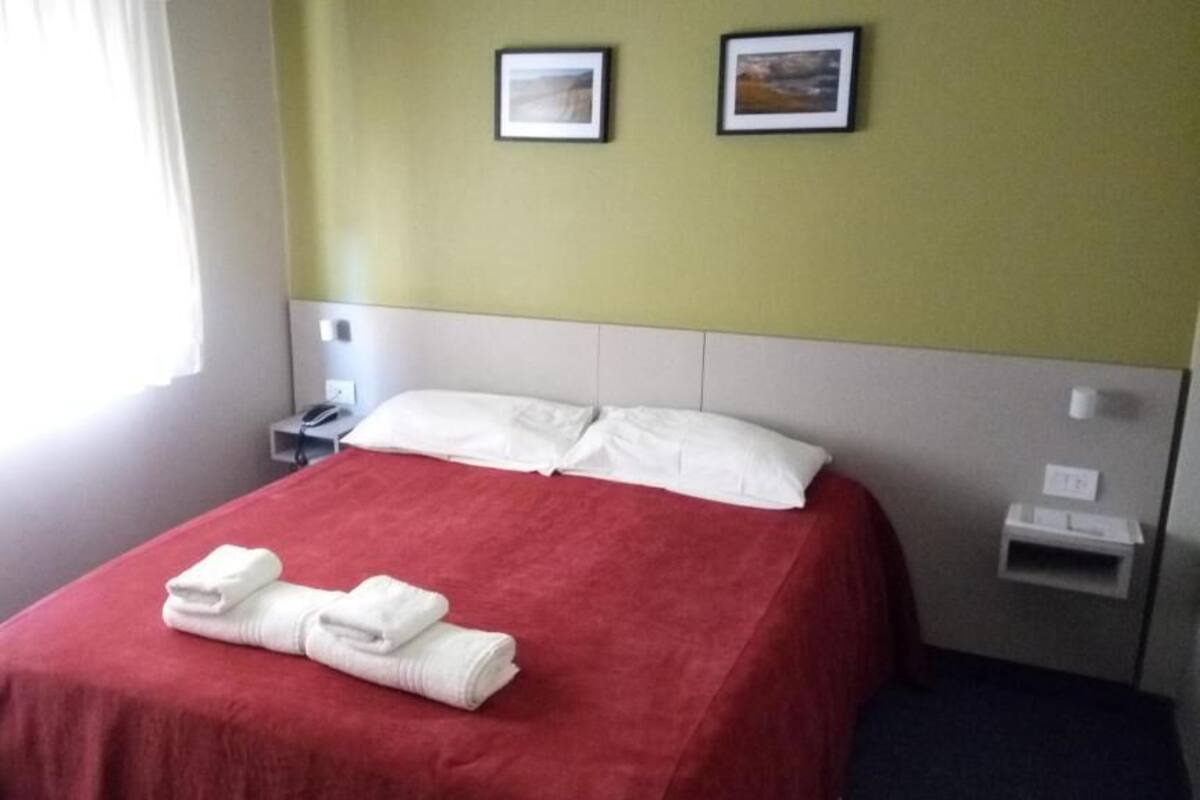 Une chambre d’hôtel avec un lit, deux tables de chevet, une petite table et une fenêtre avec des rideaux.