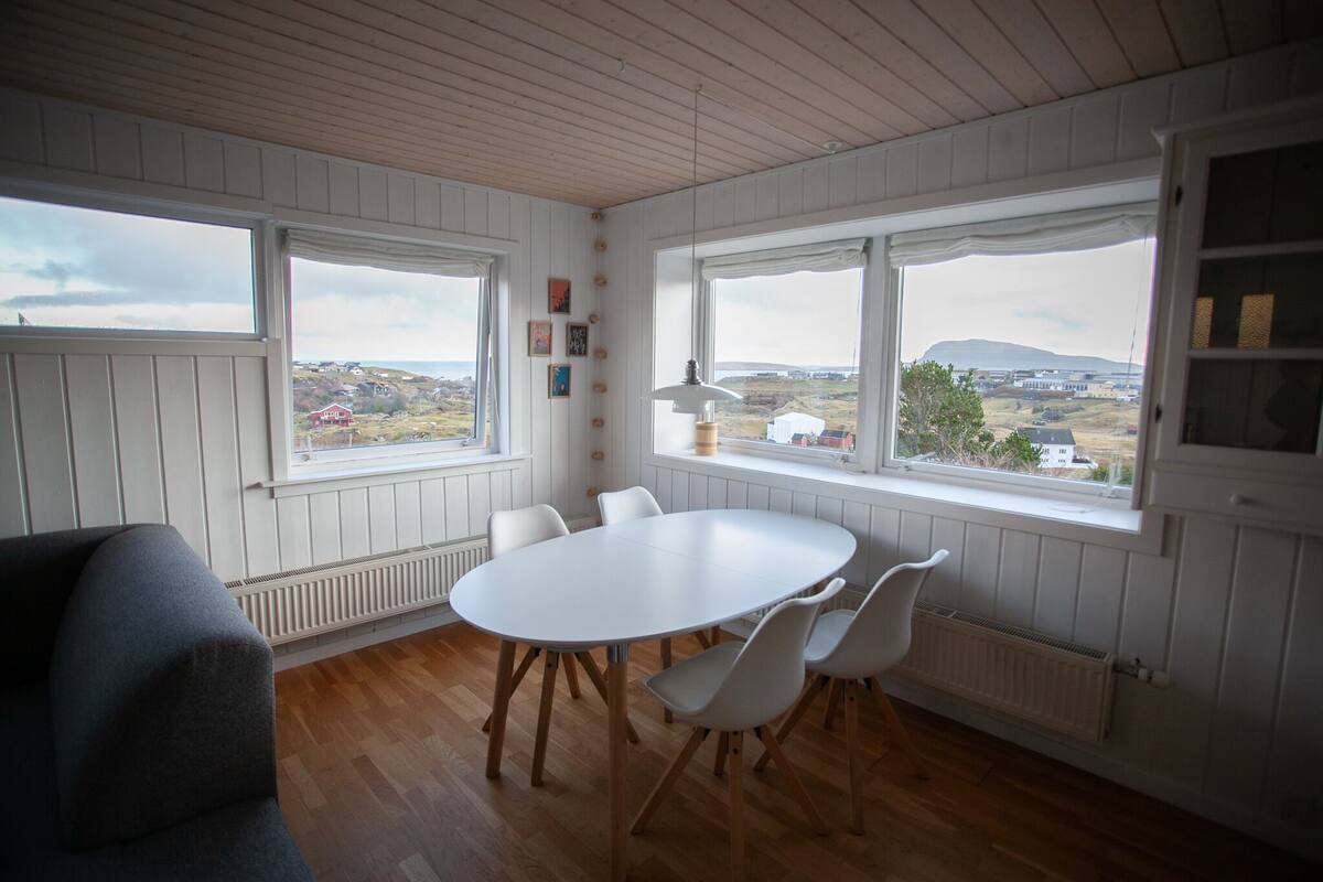 Una habitación con una mesa y sillas blancas, ventanas amplias con vistas a un paisaje, y paneles de madera en las paredes.