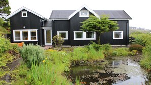 Una casa de dos pisos con fachada negra y detalles blancos, rodeada por un paisaje natural con un estanque y vegetación.