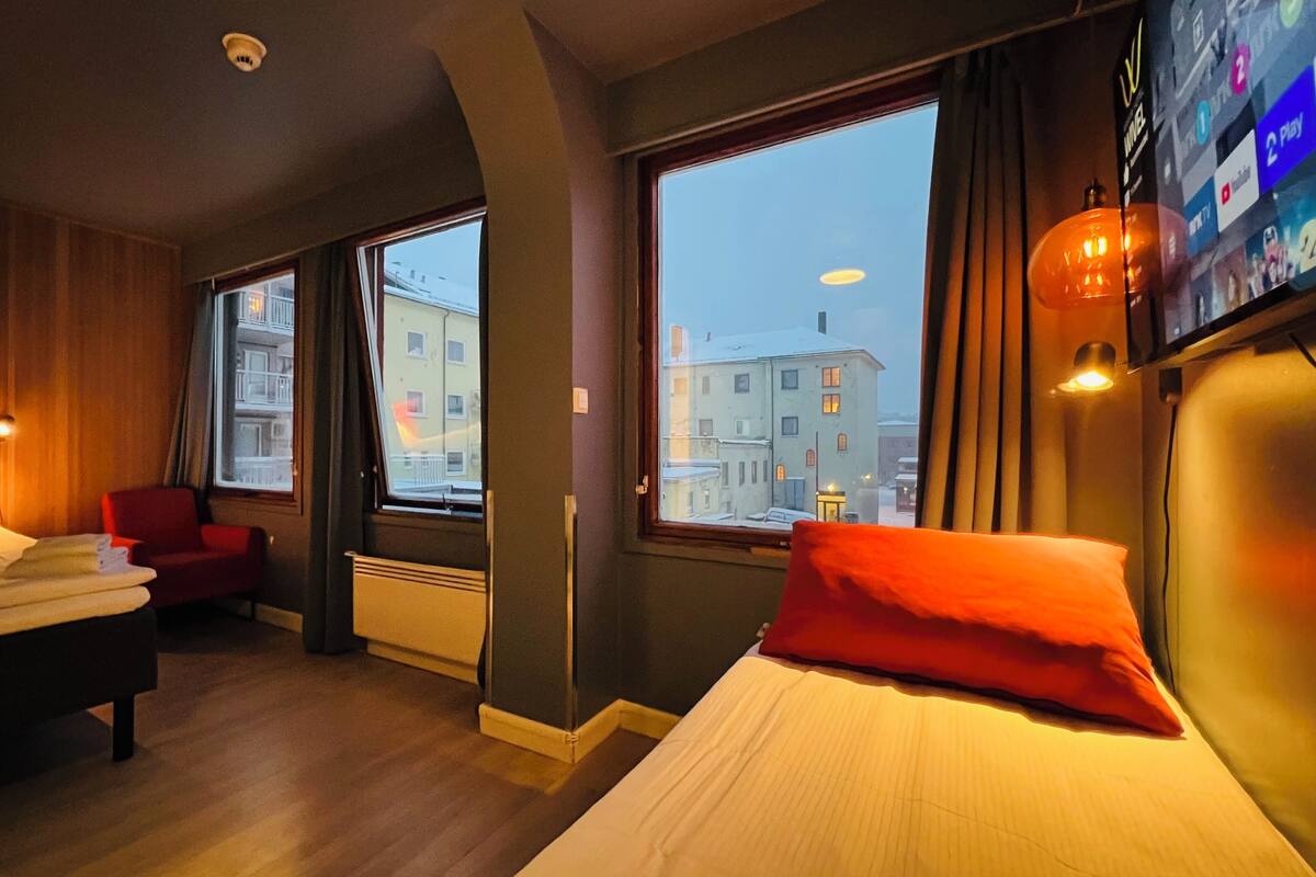A hotel room with two beds, a red chair, and large windows offering a city view.