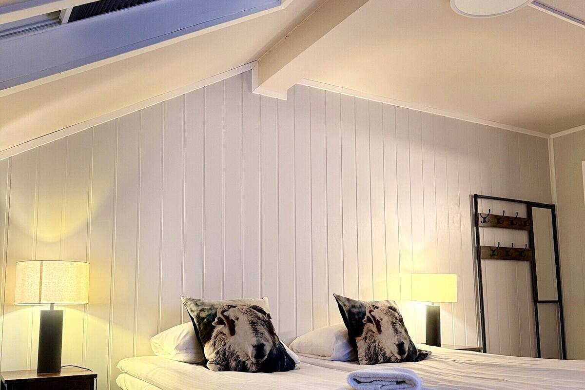 A bedroom with a bed, two pillows, a nightstand with a lamp, a ceiling light, and a coat rack.
