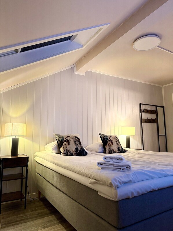 A bedroom with a bed, two pillows, a nightstand with a lamp, a ceiling light, and a coat rack. - Narvik Hotel Wivel (Narvik)