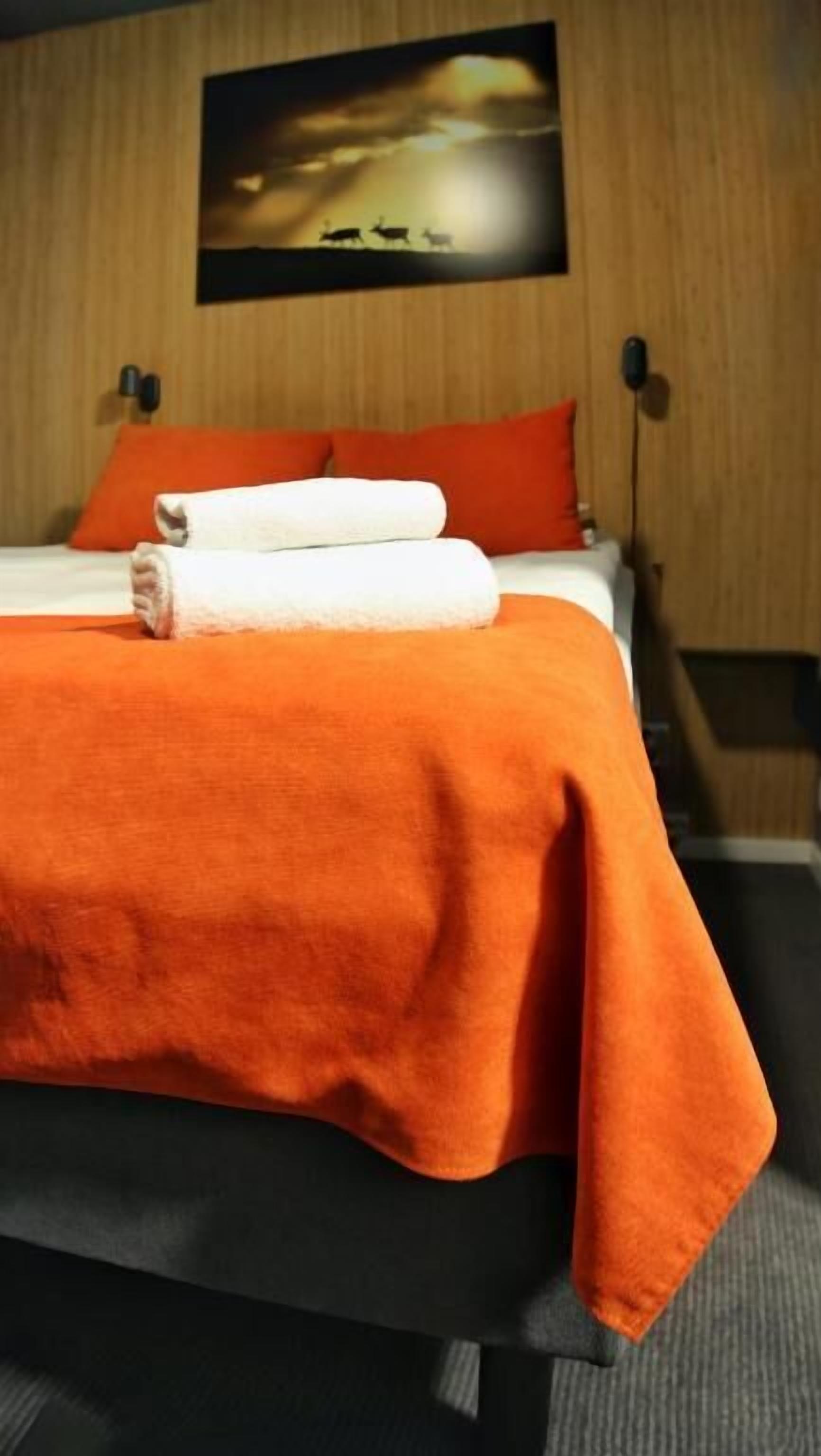 A hotel room with a bed, orange bedding, a painting on the wall, and a wooden headboard.