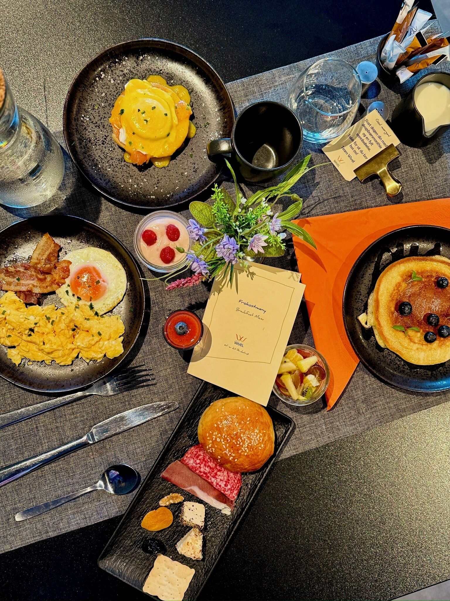 A breakfast spread with scrambled eggs, bacon, pancakes, and various pastries on a table.