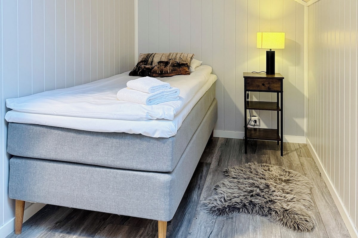 A small, neatly arranged bedroom with a single bed, a bedside table with a lamp, and a fluffy rug on the floor.
