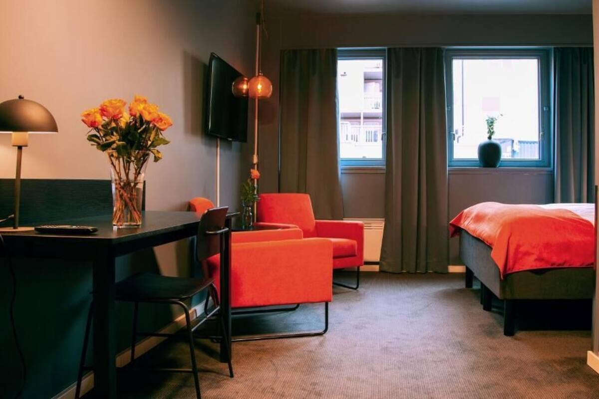 A hotel room with a desk, two chairs, a bed, a TV, and a window with curtains.