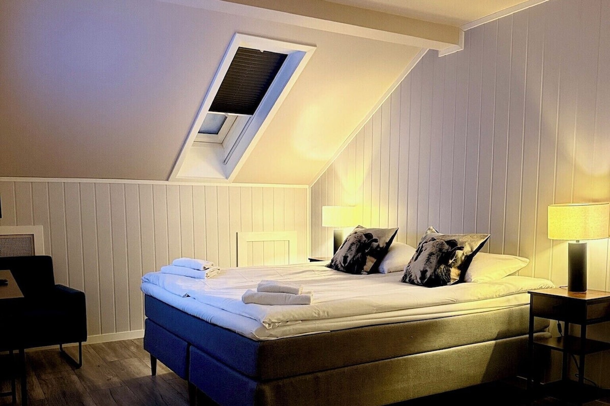 A bedroom with a sloped ceiling, a skylight, a bed with pillows, a bedside table with a lamp, and a chair.