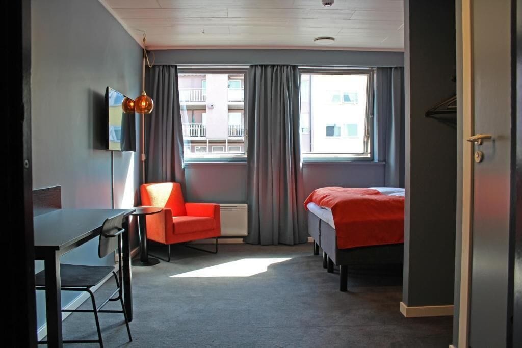 A small hotel room with a single bed, a desk, a chair, and a large window with curtains.