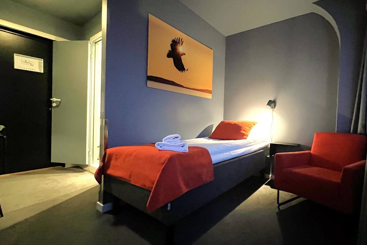 A hotel room with a bed, a chair, a nightstand, and a wall-mounted lamp.