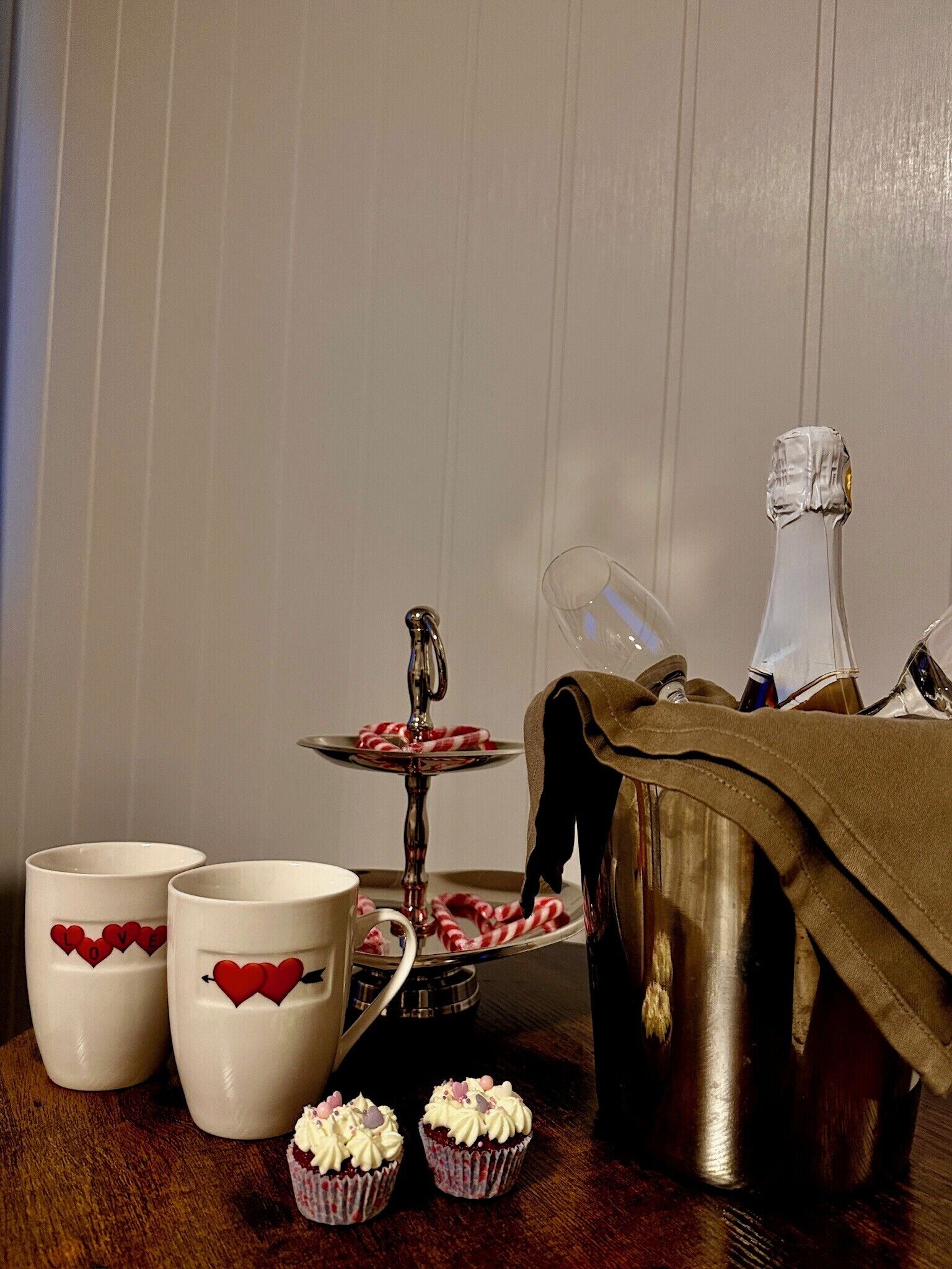 A table with two white mugs, a small cake stand with candy canes, a bottle of champagne, and two cupcakes.