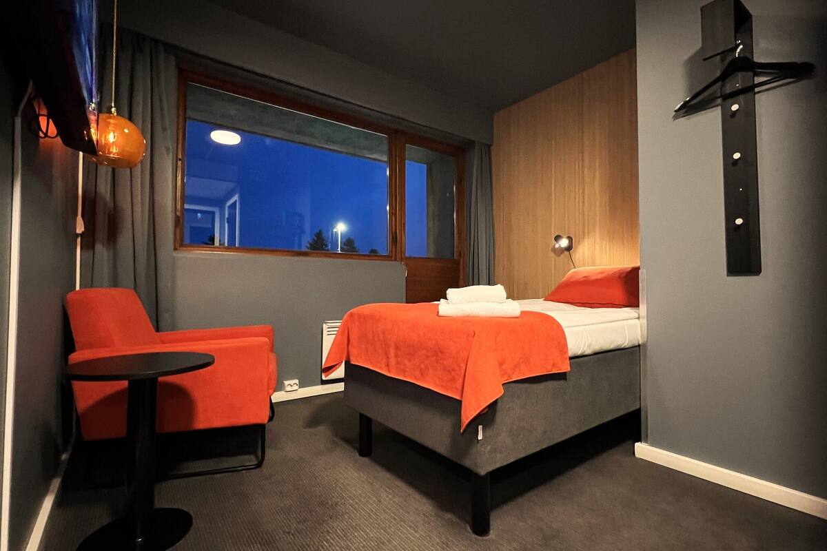 A hotel room with a bed, a chair, a small table, a window, and a wall-mounted light fixture.