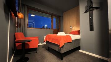 Habitación de hotel con cama, una silla, una mesita, ventana y una lámpara fijada a la pared.