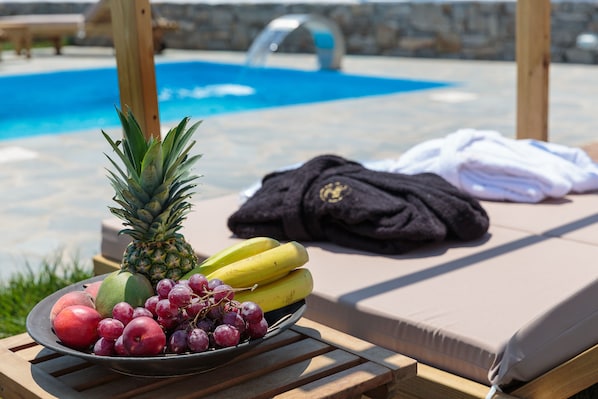 A poolside lounge with a fruit bowl containing pineapple, bananas, apples, and grapes, and a folded towel on a cushioned chair.