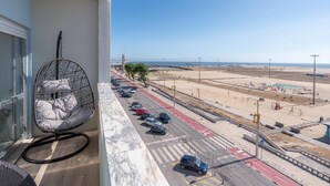 A balcony with a hanging chair, a view of a beach, a parking lot, and a road with a pedestrian crossing.