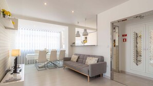 A modern living room with a grey sofa, a dining table with chairs, and a desk with a lamp.