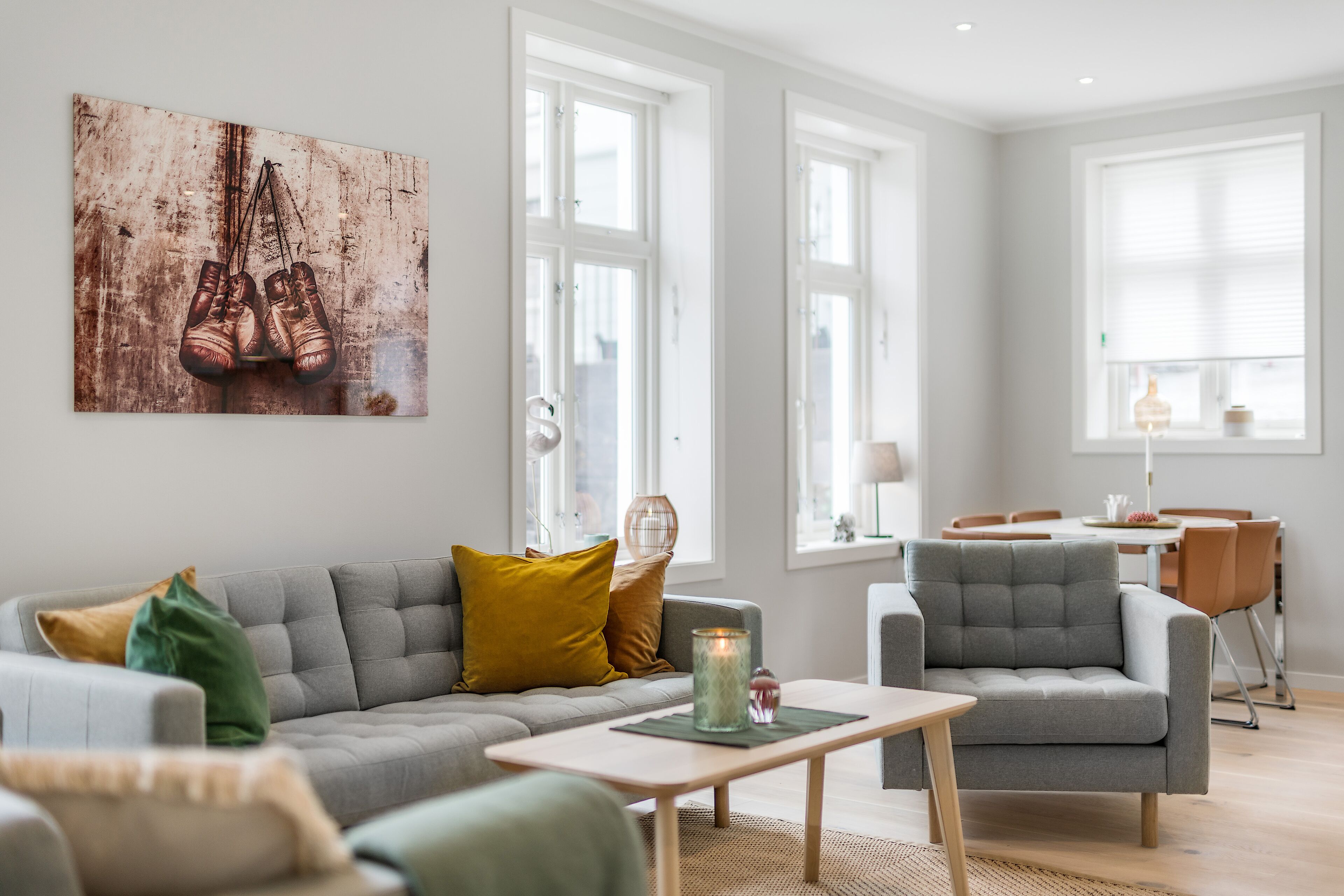 A modern living room with a grey sofa, a small table, and a chair. There is a painting of shoes on the wall.