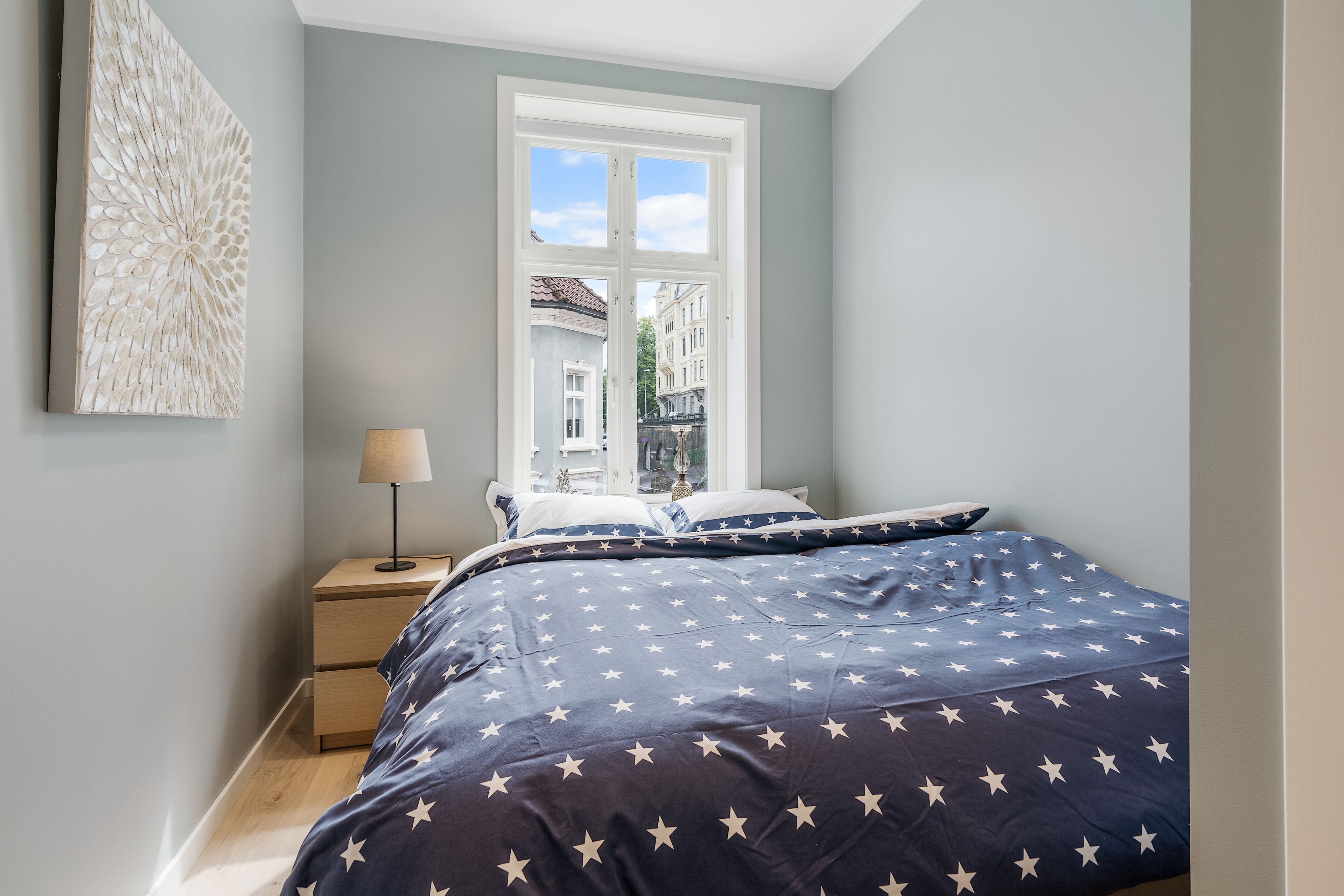 A bedroom with a bed, a nightstand, a lamp, and a window with a view of buildings.