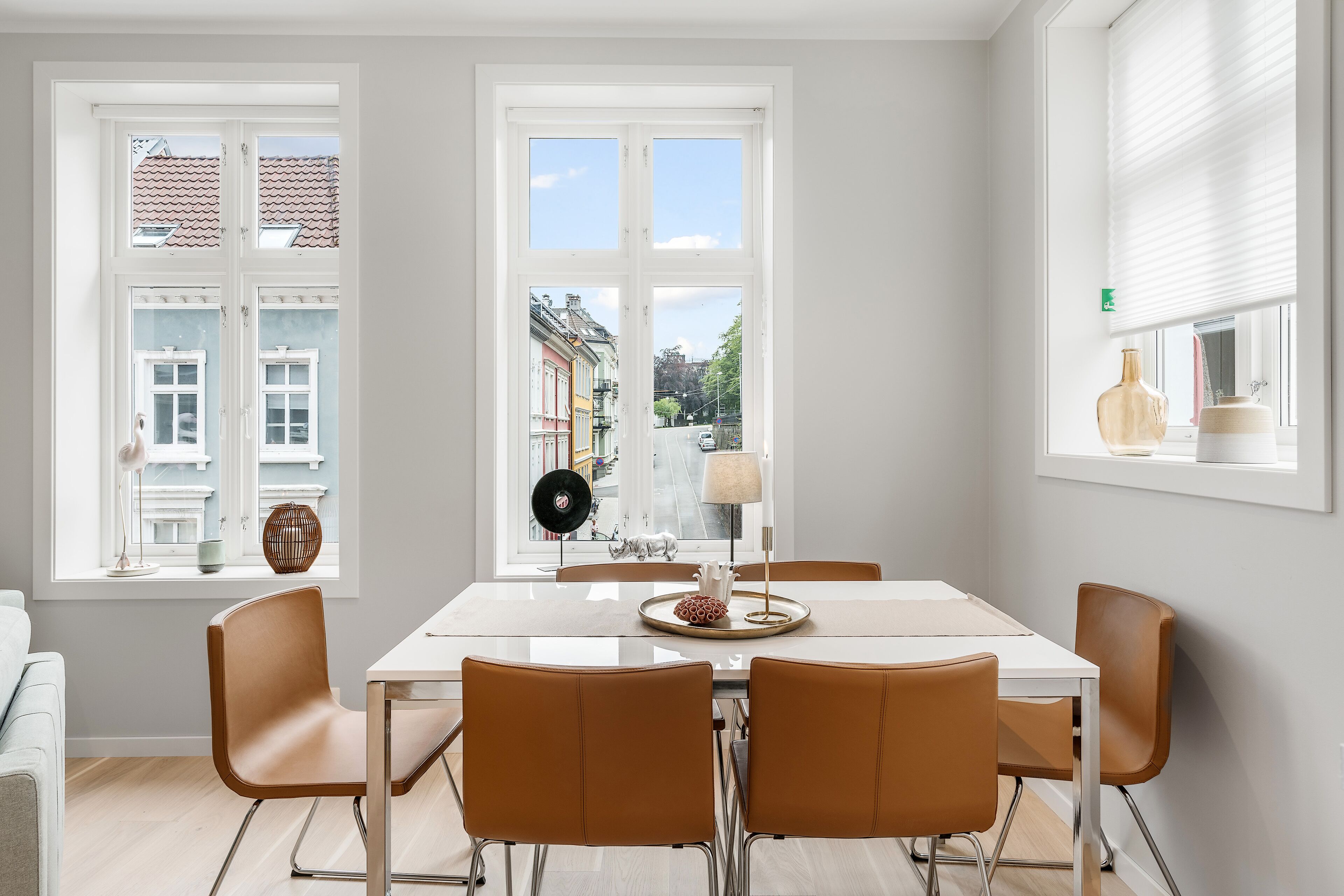 A dining area with a white table and four brown chairs, large windows with a view of buildings, and a vase with flowers on the windowsill.