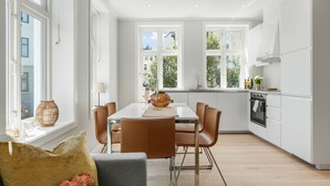 A modern kitchen with white cabinets, a dining area with a white table and brown chairs, and large windows providing natural light. - Barfot Apartments (Bergen)