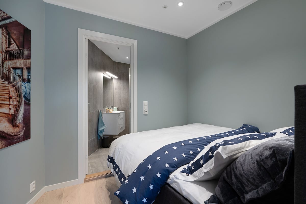 A bedroom with a bed, a painting on the wall, and a bathroom visible through an open door.