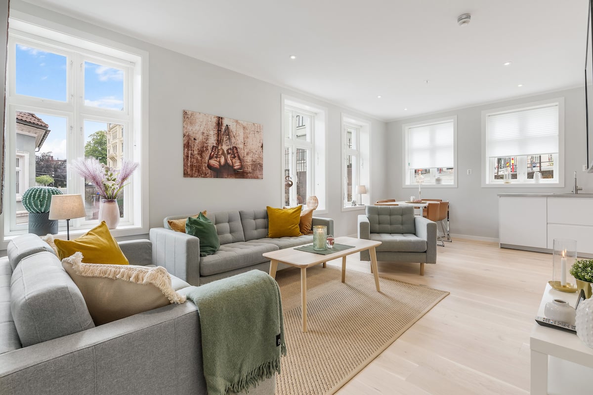 A spacious living room with a grey sofa, a small table, and a coffee table. There is a painting on the wall and a window with white blinds.