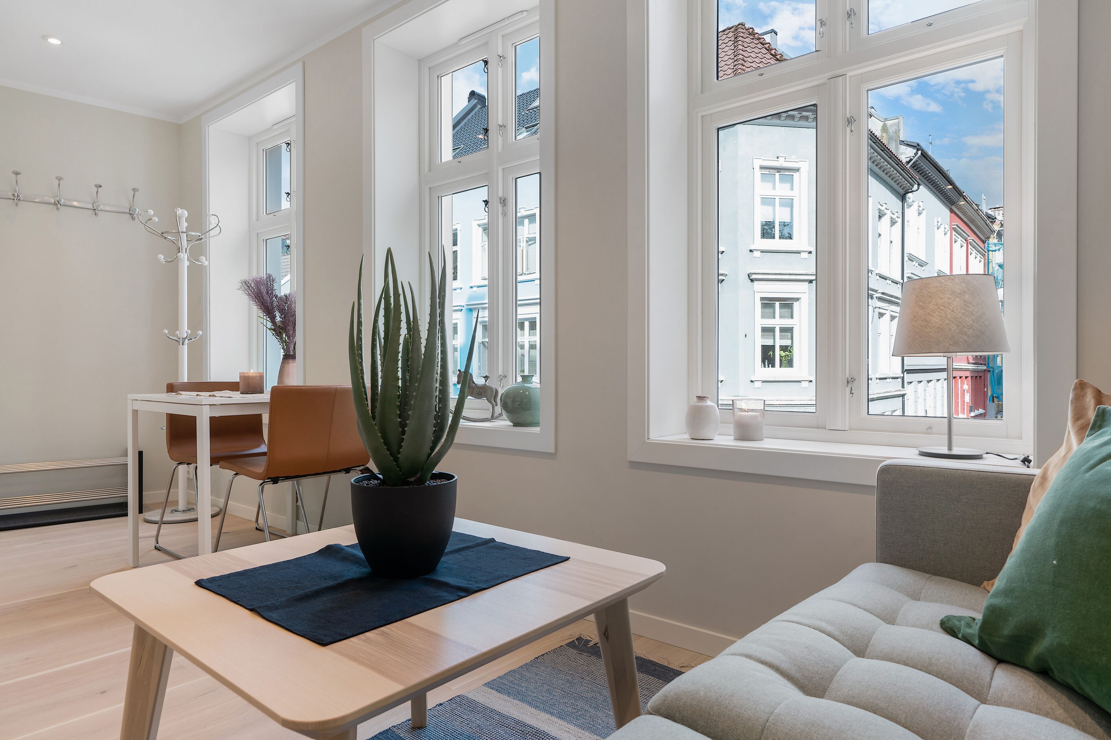 A modern living room with a sofa, a coffee table, a dining table with chairs, a potted plant, and large windows offering a view of the cityscape.
