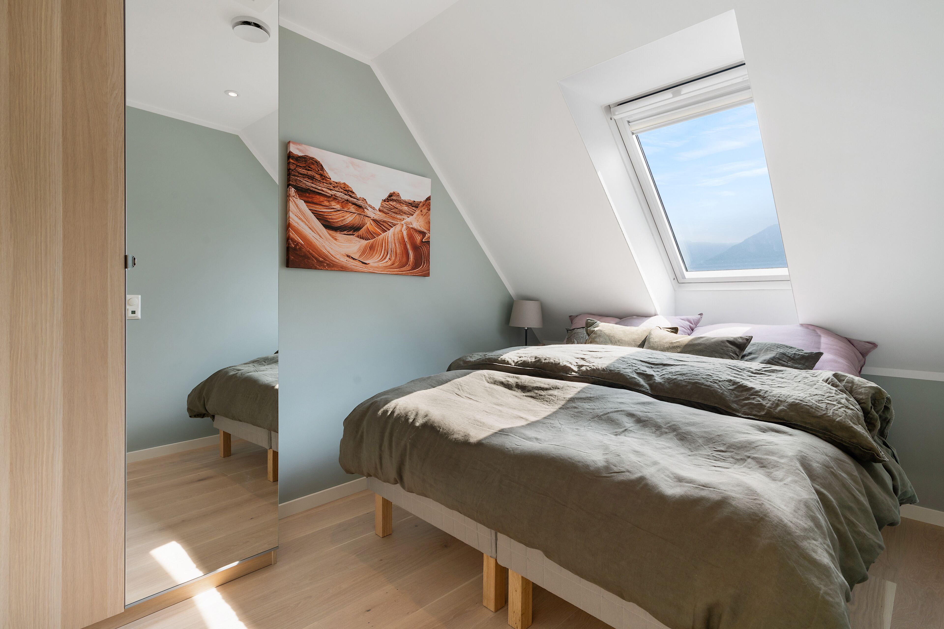Een slaapkamer met een schuine kap, dakraam, een bed met een beige en bruine dekbedovertrek, en een schilderij van een kloof aan de muur.