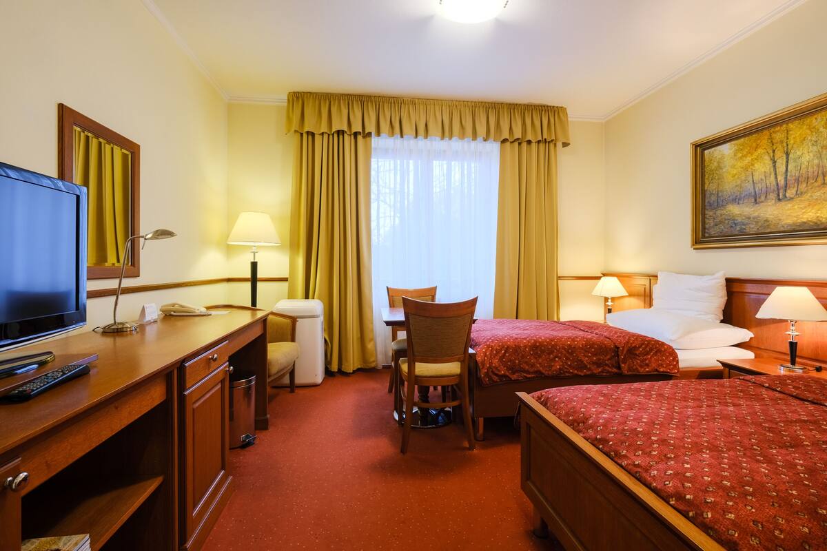 A hotel room with a bed, a desk with a television, a chair, a small table, and a window with curtains.